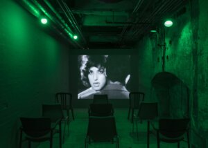 A black-and-white video projection showing a closeup of an actress at the far end of green-cast basement space.