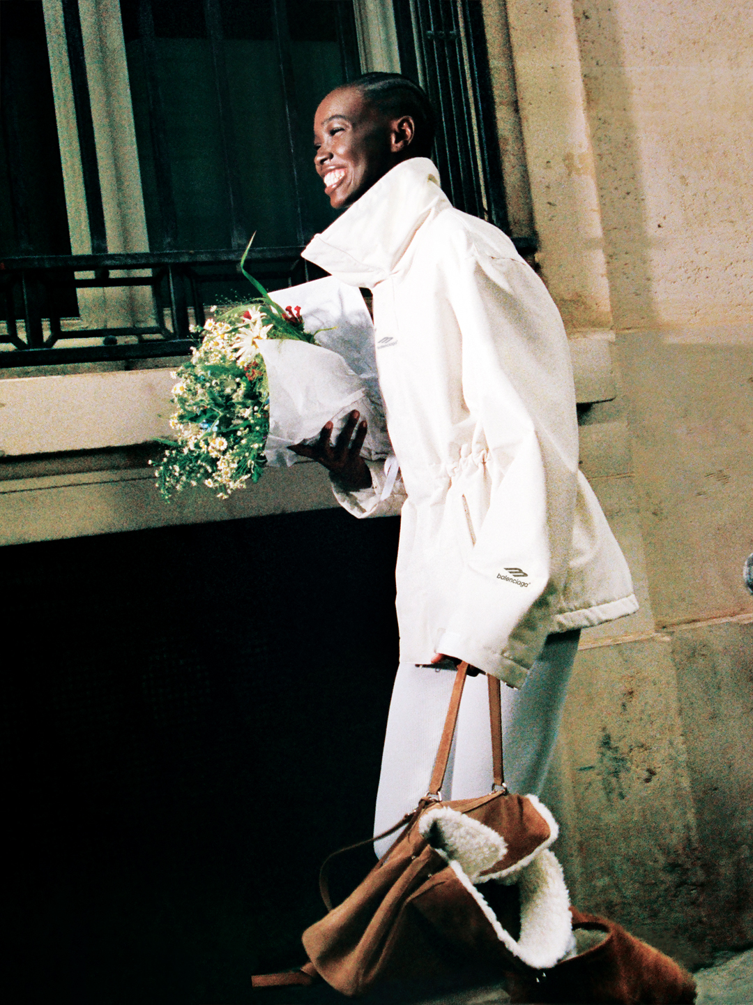 Portrait of a woman wearing the Balenciaga ski collection white nylon shell jacket, carrying a shearling lined Rodeo bag as well as a bouquet of flowers