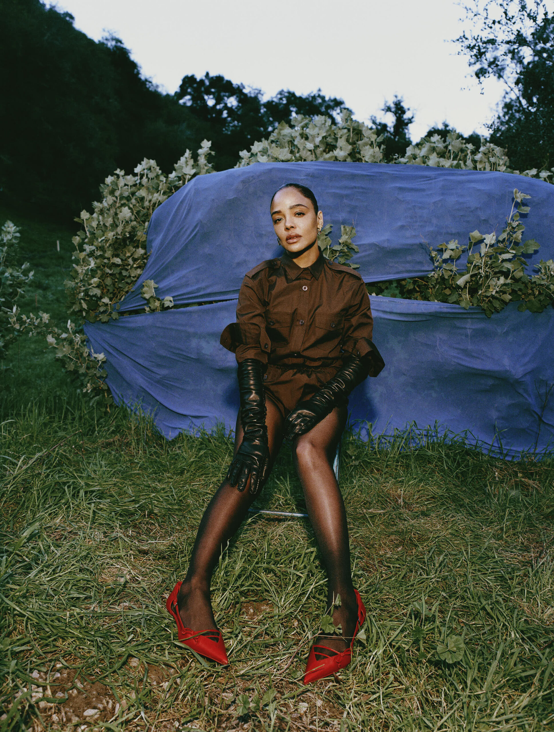 Actress Tessa Thompson poses outside in a field wearing a brown shirt dress and red heels.