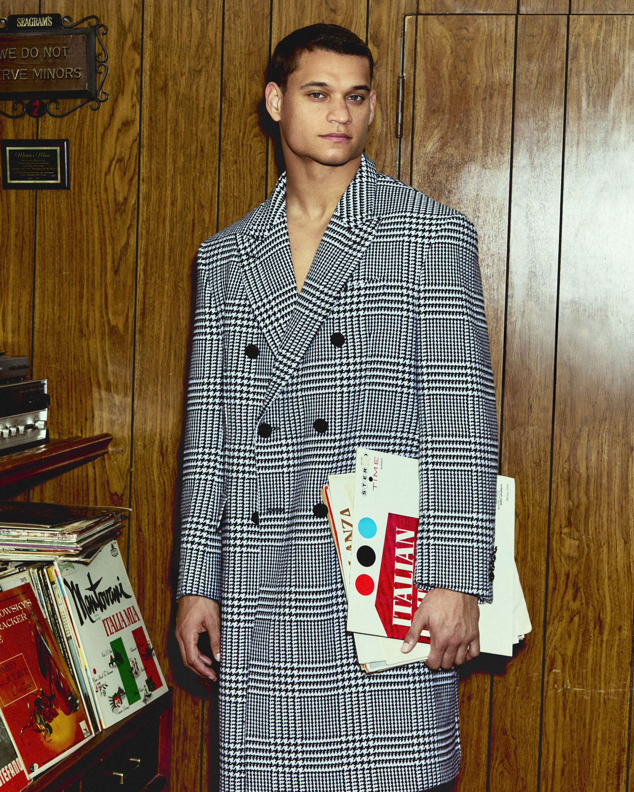 Tyriq Withers poses in a wood paneled room wearing a Gucci Prince of Wales wool trench coat.