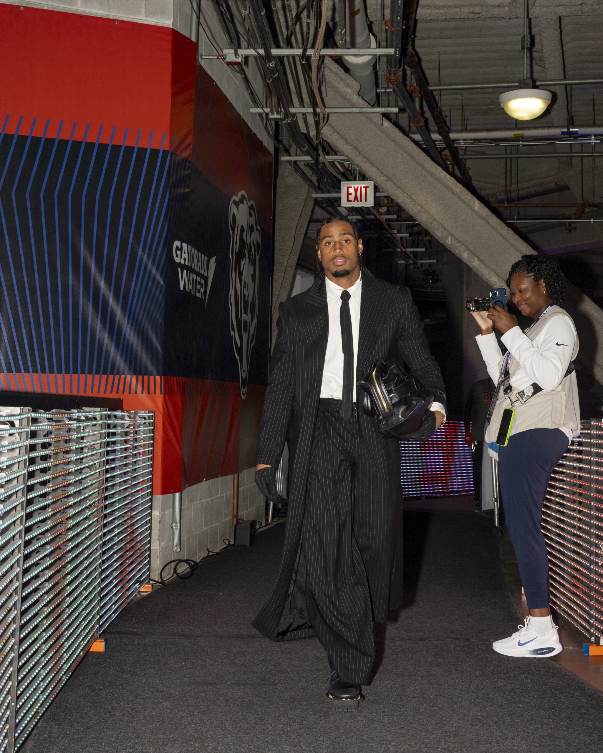 Kyler Gordon of the Chicago Bears in a black coat, black tie, white shirt, leather bag, and black shoes walking through the tunnel