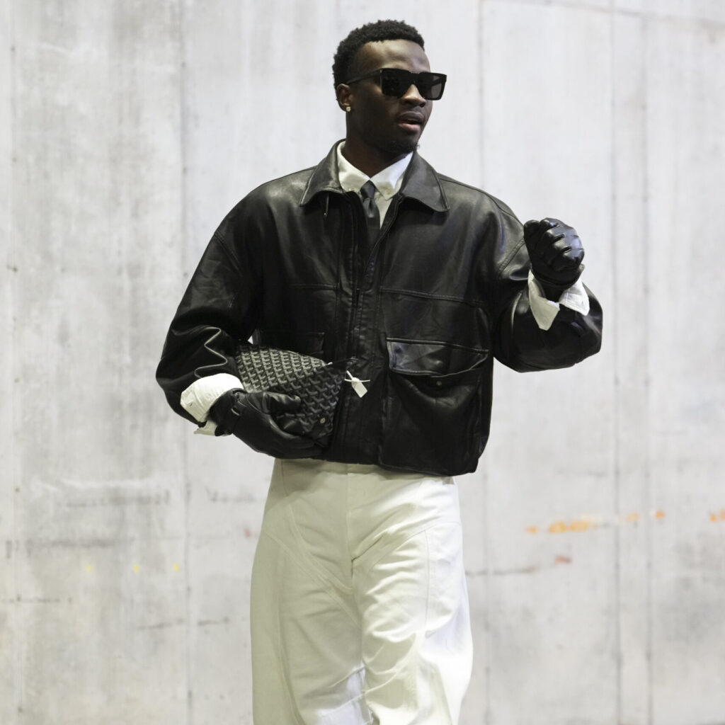 Mike Sainristil of the Washington Commanders in a leather jacket and gloves, sunglasses, black tie, white button-down, white slacks, and black sneakers in his tunnel look