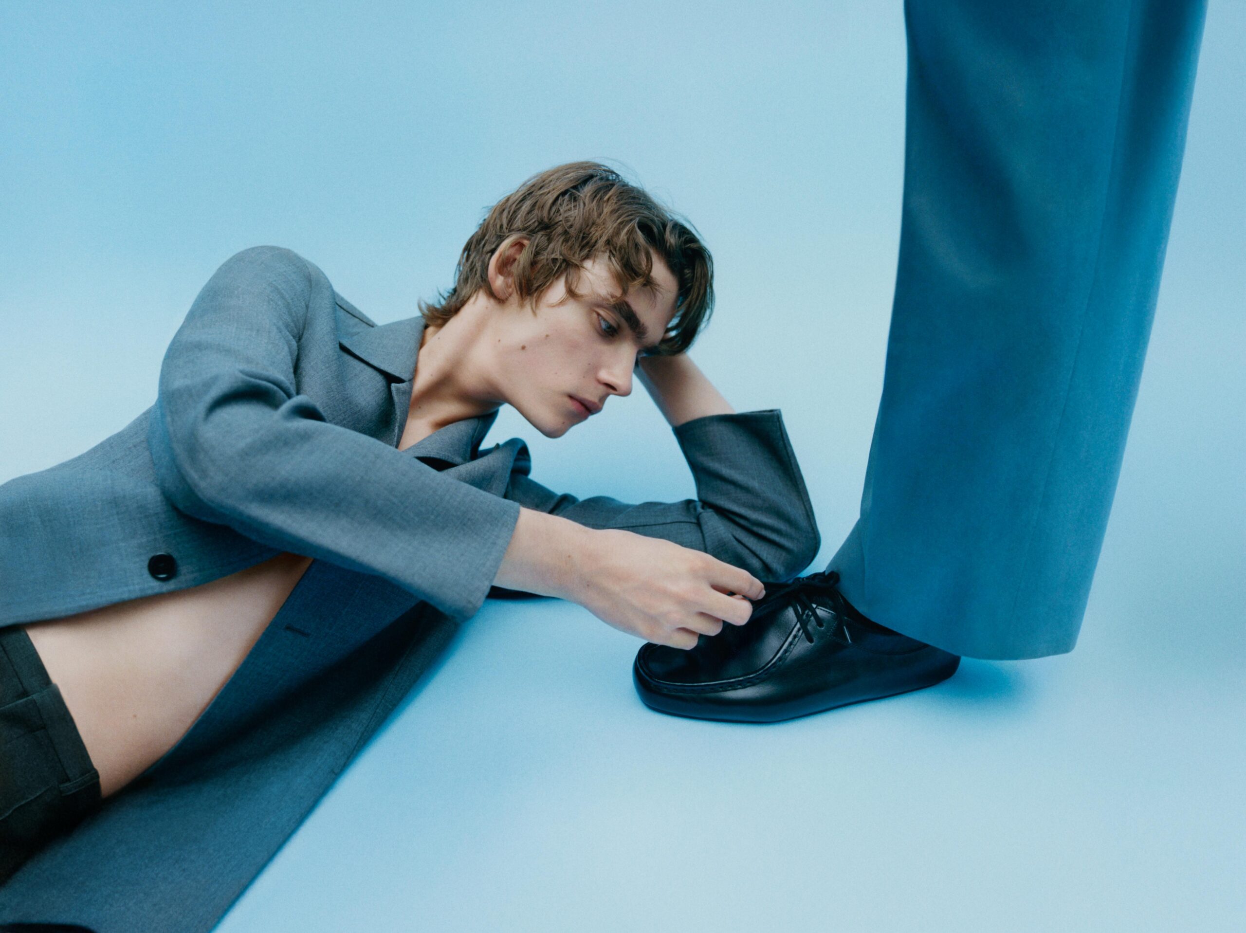 A model plays with a peer's shoe in the Jil Sander Spring/Summer 2026 campaign against a blue background