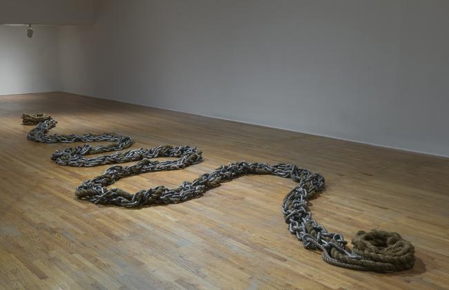 A sculpture made of steel chains and rope configured like a long snake on a wood floor. 